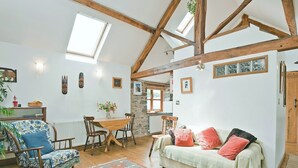 Uma sala de estar acolhedora, com teto alto, vigas de madeira aparente, área de jantar com mesa e cadeiras, e um sofá com almofadas.