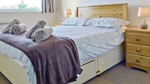 A neatly made bed with rolled towels, a bedside table with lamps, and a window with curtains. - 3 bedroom accommodation in Fairbourne (Fairbourne)