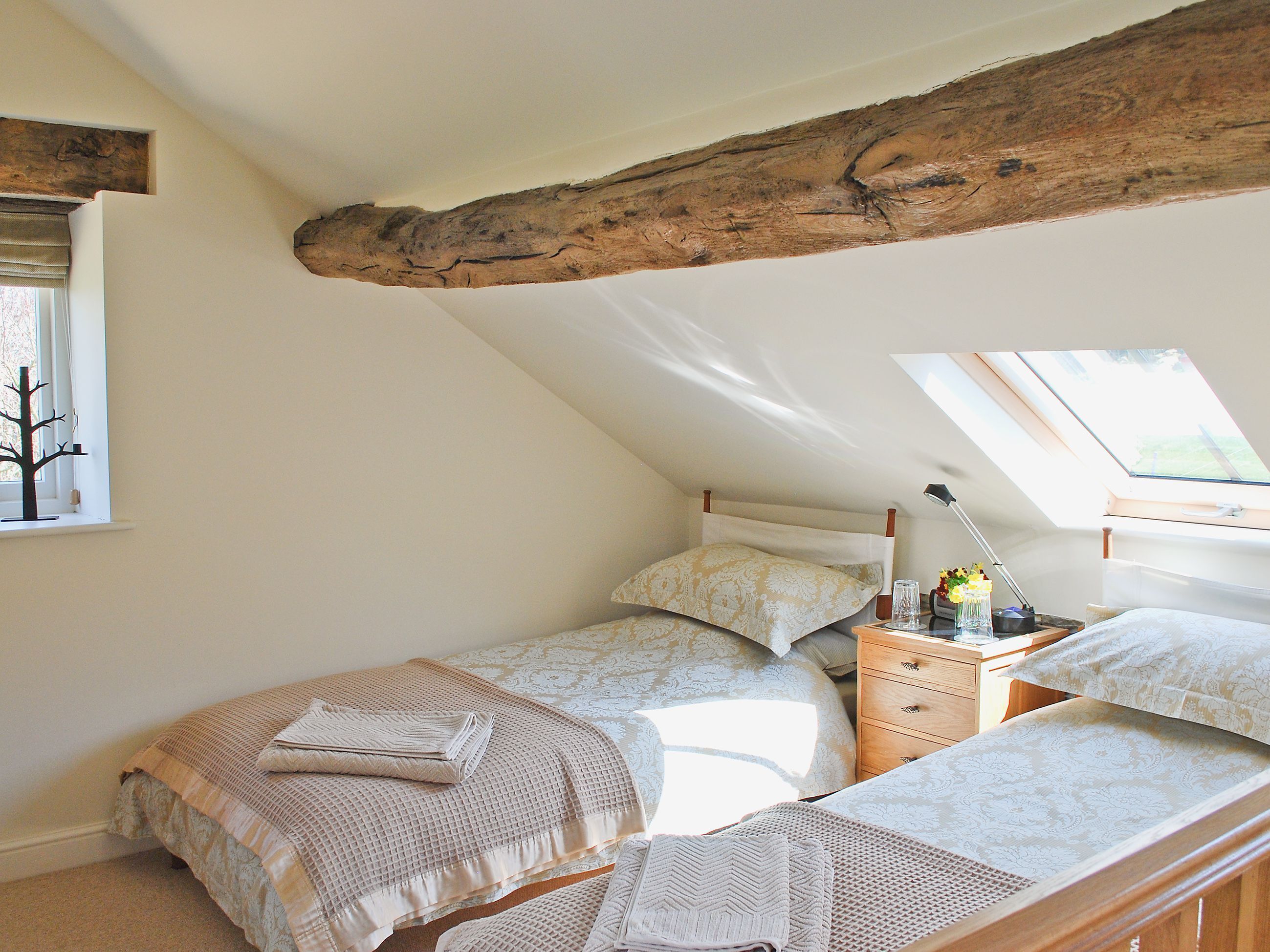 A bedroom with a sloped ceiling, a bed with a patterned bedspread, a bedside table with a lamp, and a window with a view of the outdoors.