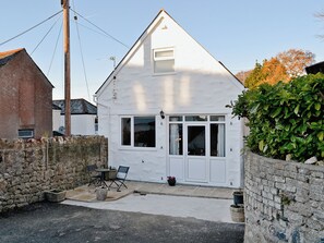 A white two-story house with a stone wall, a small patio with chairs and a table, and a large bush in front.