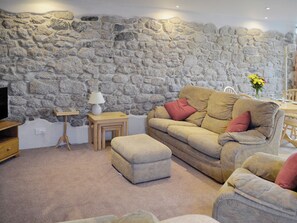 Un salon avec un mur en pierre, un canapé, un pouf, une petite table avec une lampe et un téléviseur.