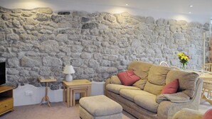 A living room with a stone wall, a beige sofa, an ottoman, a small wooden table with a lamp, and a TV on a wooden stand. - 1 bedroom accommodation in St Austell (St Austell)