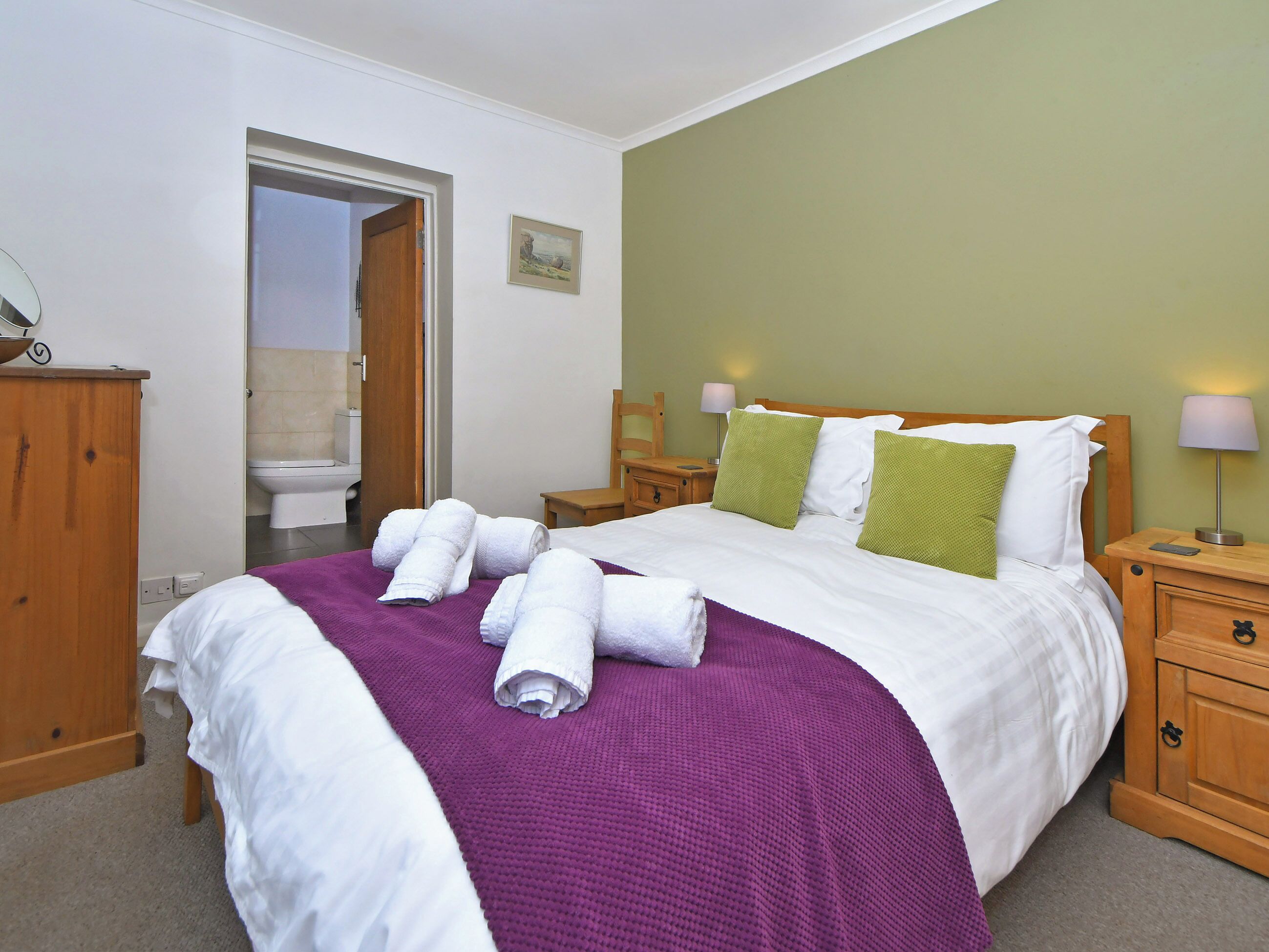 A neatly made bed with white and green pillows, a purple blanket, and rolled white towels. There is a wooden nightstand with a lamp, a wooden dresser, and a bathroom visible through an open door.