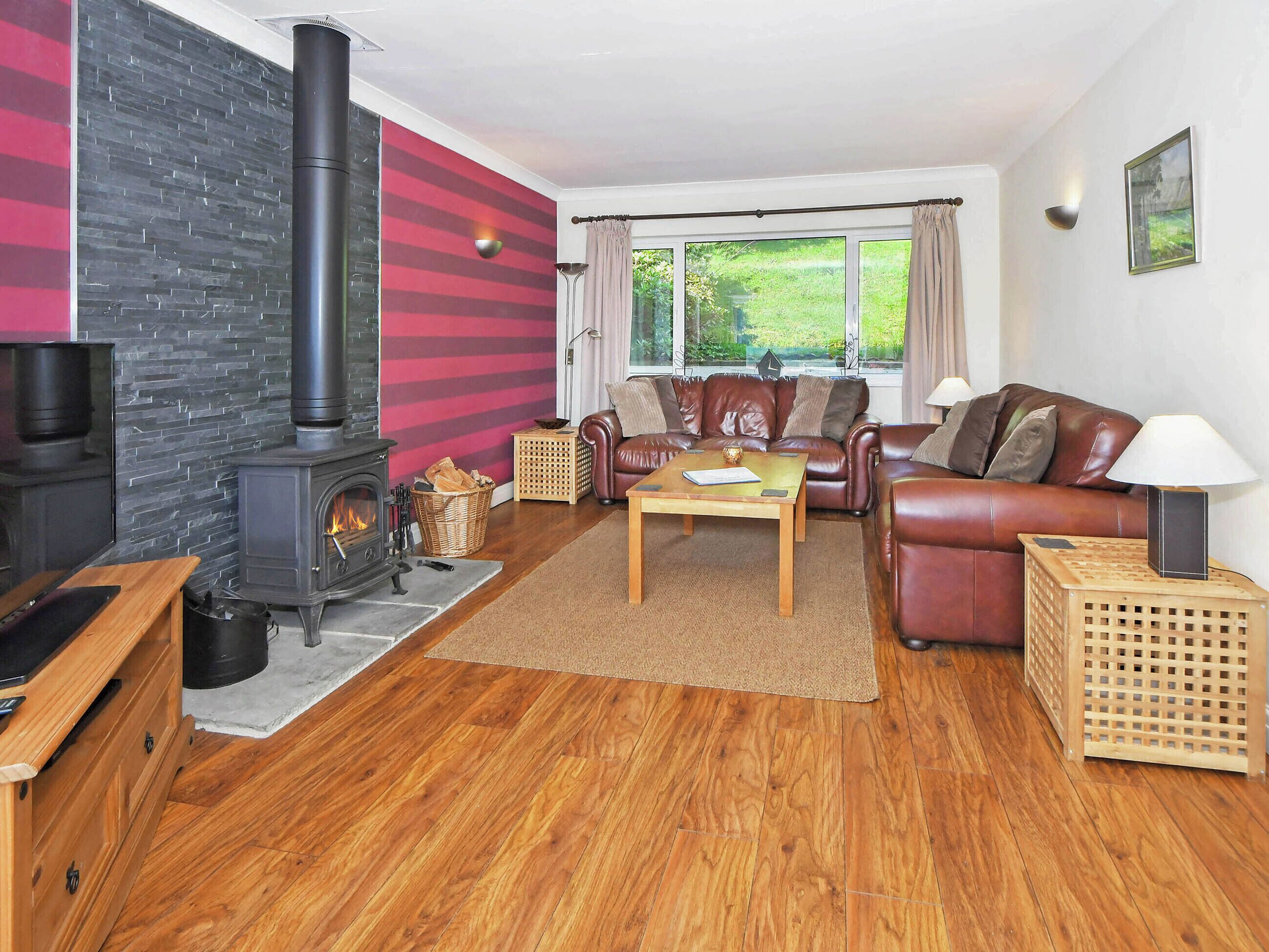 A cozy living room with a fireplace, leather sofa, wooden coffee table, and a view of greenery outside.