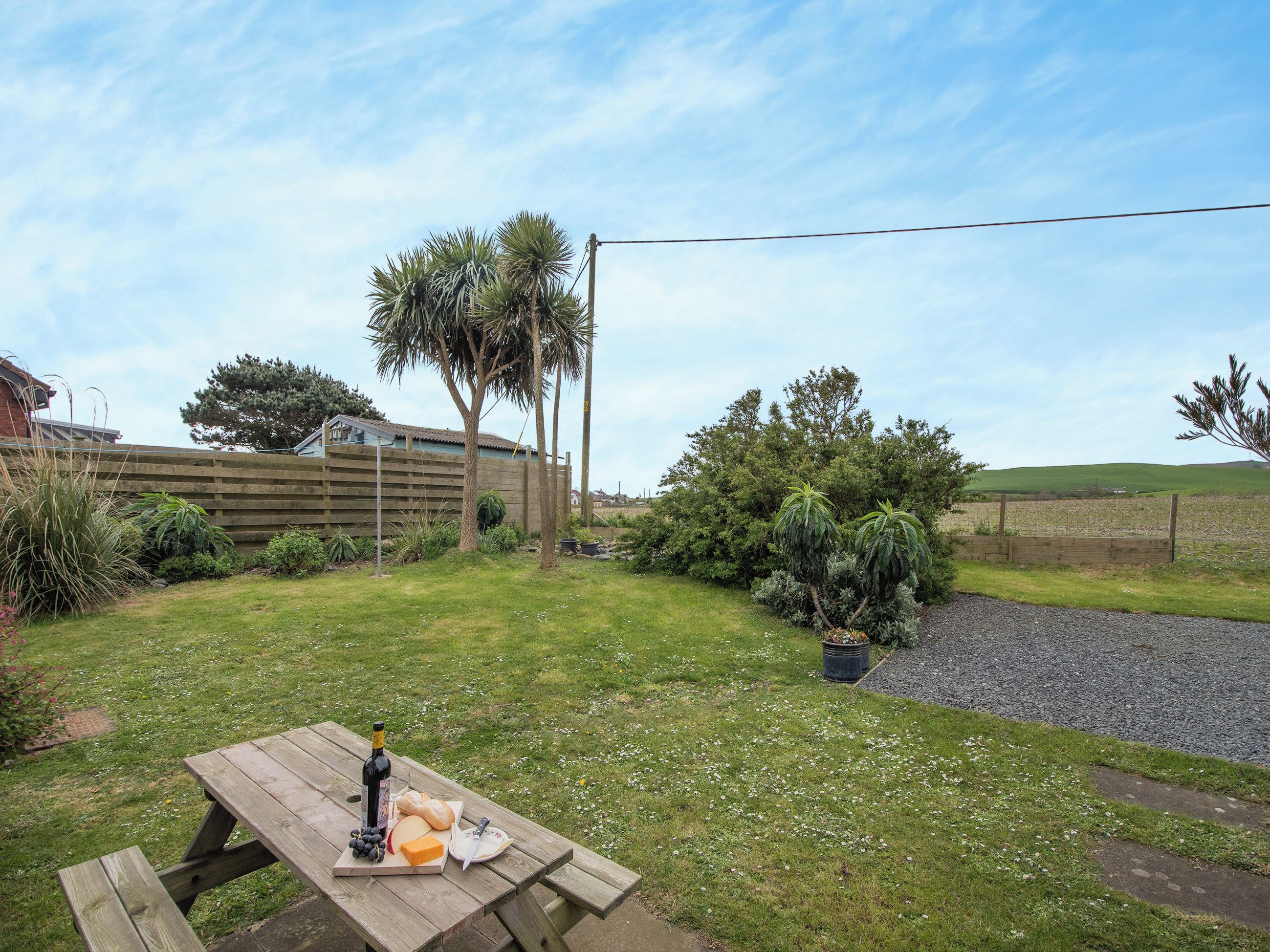A picnic table with food and a bottle, surrounded by a grassy yard with trees and a fence.