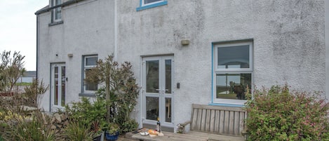Una casa de dos pisos con un patio jardín, un banco de madera y una mesa con una botella y pan.