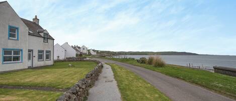 A coastal road with stone walls and a grassy path leading to houses with blue trim.