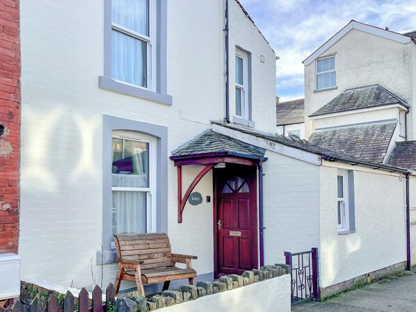 A white building with a red door, a wooden bench outside, and a stone wall.