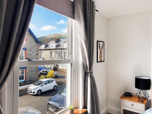 A room with a large window, a white bedside table, a lamp, and a framed picture on the wall. - 2 bedroom accommodation in Keswick (Keswick)