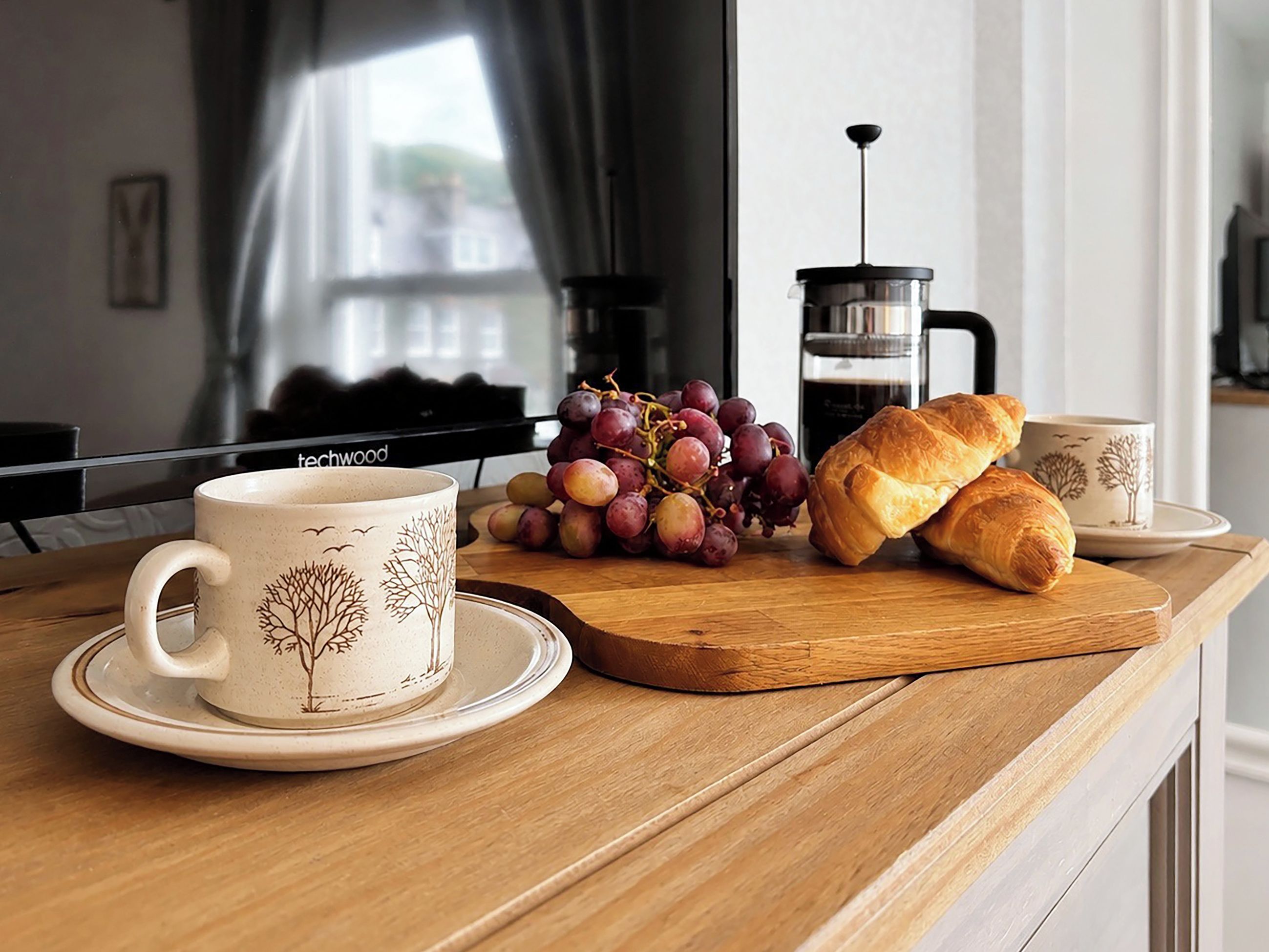 Une table en bois avec une machine à café, une tasse, une assiette de raisins et des croissants.