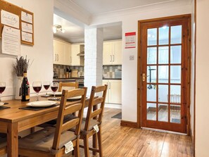 Comedor con una mesa de madera dispuesta para cuatro personas, una botella de vino y copas sobre la mesa. Se observan armarios blancos y un pilar de ladrillo al fondo. Se ve una puerta con paneles de vidrio a la derecha.