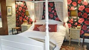 A four-poster canopy bed with white bedding, a floral wallpaper background, and two bedside tables with lamps.