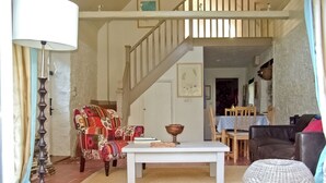 A living room with a staircase, a patterned armchair, a white table, and a wicker ottoman.