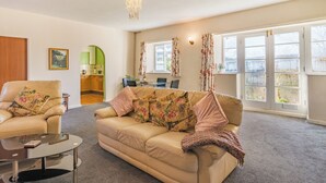 Ein Wohnzimmer mit einem beigefarbenen Sofa, einem Glastisch und einem Kronleuchter. Es gibt einen Essbereich mit Tisch und Stühlen, und durch einen Bogen ist eine Küche zu sehen.