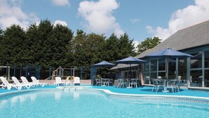 Ein Schwimmbecken mit Liegestühlen und Sonnenschirmen, umgeben von Bäumen und einem Gebäude mit großen Fenstern.