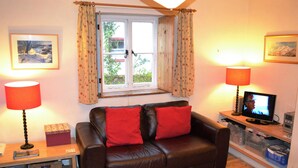 Un salon chaleureux avec un canapé en cuir marron, deux coussins rouges, une table d’appoint en bois, une lampe, un téléviseur et une fenêtre avec des rideaux.