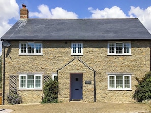 Une petite maison en pierre avec un toit sombre, deux fenêtres à l’étage, une porte centrale et une enseigne indiquant « Woodbrook Cottage ».
