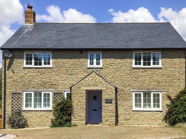 A stone cottage with a dark roof, two windows on the upper floor, a central door, and a sign reading 'Woodbrook Cottage.'