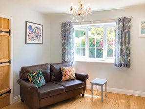 A room with a brown leather sofa, a small white table, a chandelier, a window with floral curtains, and a framed picture on the wall.