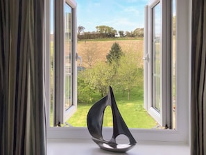 Une pièce avec vue sur le paysage, grâce à de grandes fenêtres ouvertes, une sculpture abstraite noire et un champ verdoyant parsemé d’arbres.