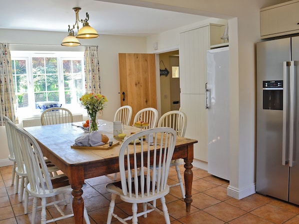 Un coin repas avec une table en bois, des chaises blanches, un vase de fleurs et une cuisine équipée d’un réfrigérateur.