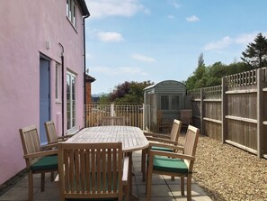 A patio with a round table and wooden chairs, a pink building, a wooden fence, and a shed in the background.