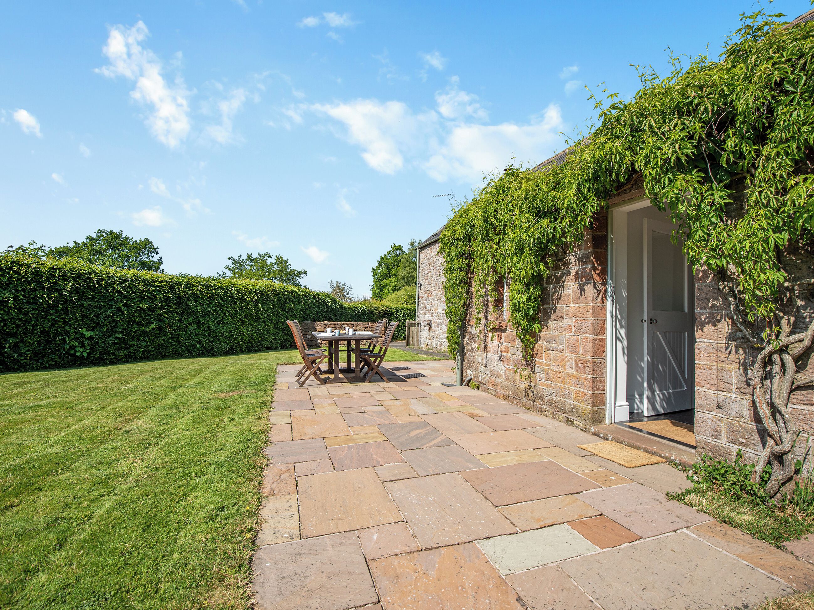A stone patio with a table and chairs, a hedge, and a stone building with a door.