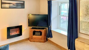 A living room with a flat-screen TV on a wooden stand, a fireplace, a window with curtains, and a framed map on the wall. - 1 bedroom accommodation in Keswick (Keswick)