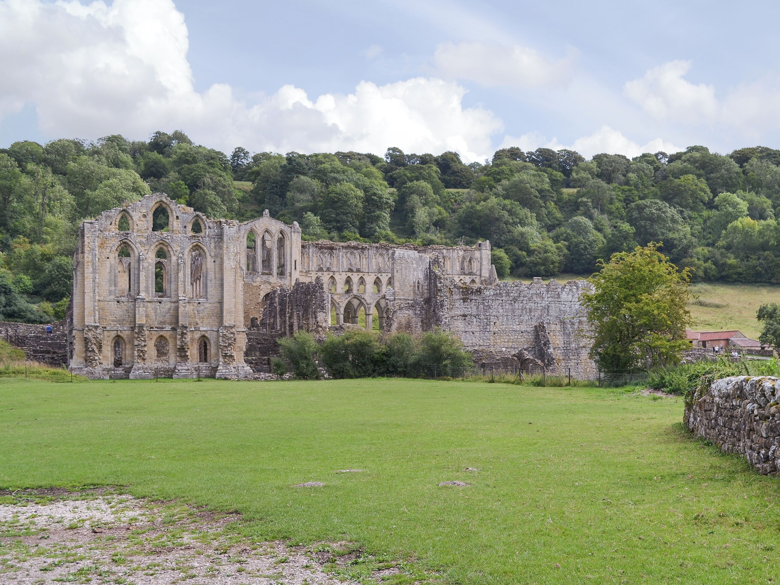 A historic stone building with arched windows and ruins, surrounded by a grassy field and dense forest.