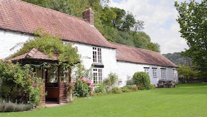 Een traditioneel wit gebouw met een rode pannendak, een houten veranda en een tuin met bloemen en struiken.