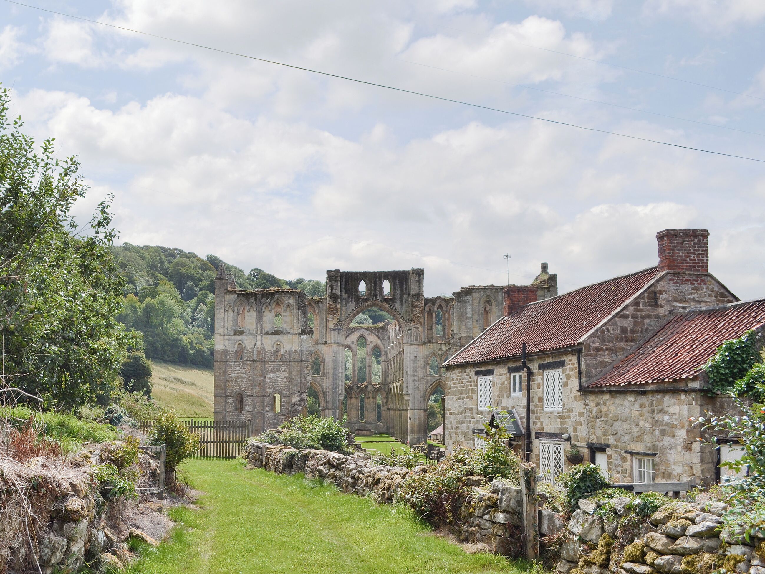 A historic stone building with a large arched entrance, a stone wall, and a grassy area.