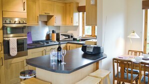 A modern kitchen with wooden cabinets, a built-in oven, a microwave, and a coffee machine. There are two wooden stools and a table with chairs.