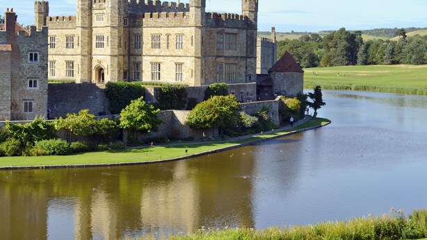 A historic castle with a moat, surrounded by lush greenery and a clear sky.