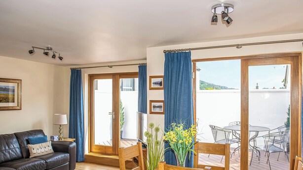 A living room with a sofa, a dining table with chairs, a TV, and a view of the outdoors. - 2 bedroom accommodation in Keswick (Keswick)