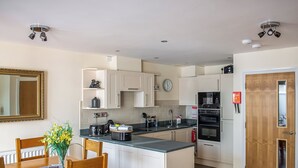 A modern kitchen with white cabinets, a built-in oven, and a coffee machine. There is a dining table with chairs and a vase of flowers. - 2 bedroom accommodation in Keswick (Keswick)