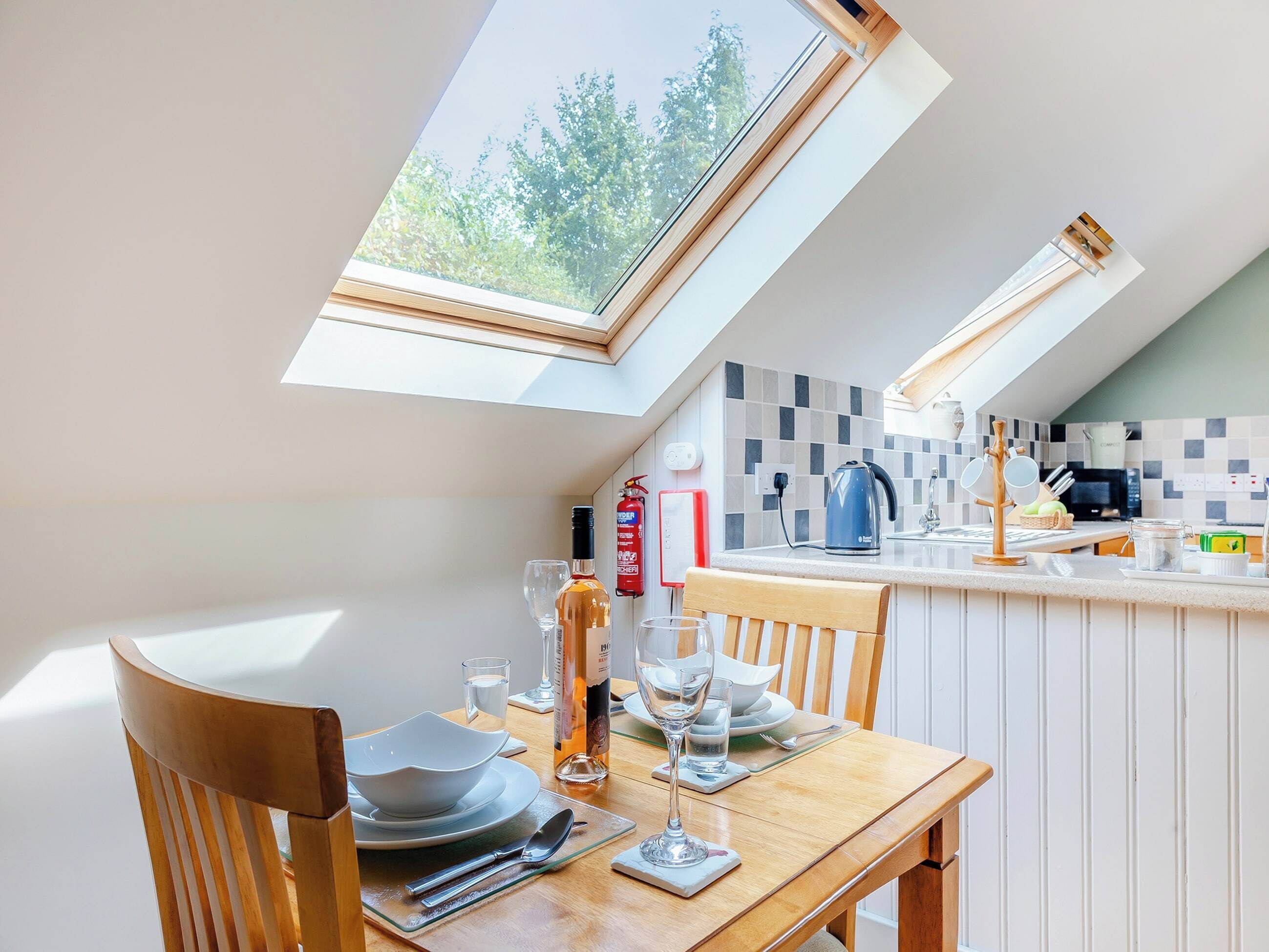 A dining area with a table set for two, a kitchen in the background, and a skylight above.