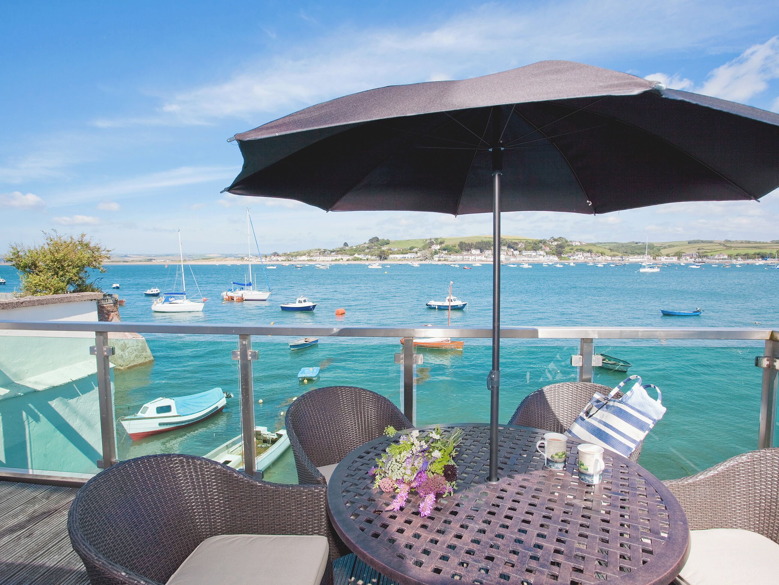 A terrace with wicker furniture, a table with a flower arrangement, and a view of boats on the water.