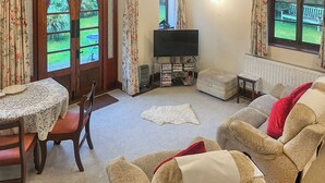 A cozy living room with a fireplace, a television, and a bookshelf. There is a dining table with chairs and a window with curtains. - 1 bedroom accommodation in Bransgore, near Christchurch (Bransgore, near Christchurch)