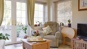 A cozy living room with a sofa, armchair, and coffee table. There are several potted plants and a television on a wooden stand.