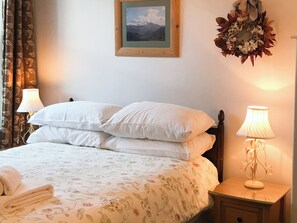 A neatly made bed with white linens and a floral-patterned duvet, a wooden nightstand with a lamp, a framed picture on the wall, and a decorative wreath. - 3 Catherine Cottages (Keswick)