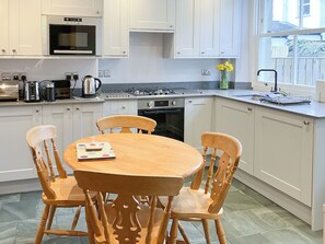 A modern kitchen with white cabinets, a wooden dining table, and chairs. There is a window with a view of the outside. - 3 Catherine Cottages (Keswick)