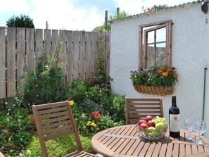 Une table en bois avec un saladier de fruits et une bouteille de vin, une chaise en bois et un panier de fleurs suspendu.
