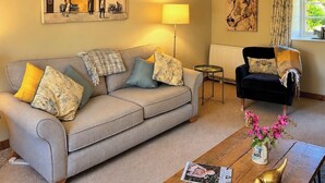 A living room with a sofa, a black armchair, a coffee table with a magazine, and a vase with flowers. - 2 bedroom accommodation in North Chideock, near Bridport (North Chideock, near Bridport)