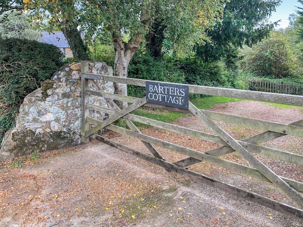 A wooden gate with a sign reading "BARTERS COTTAGE" leading to a stone wall and a pathway.
