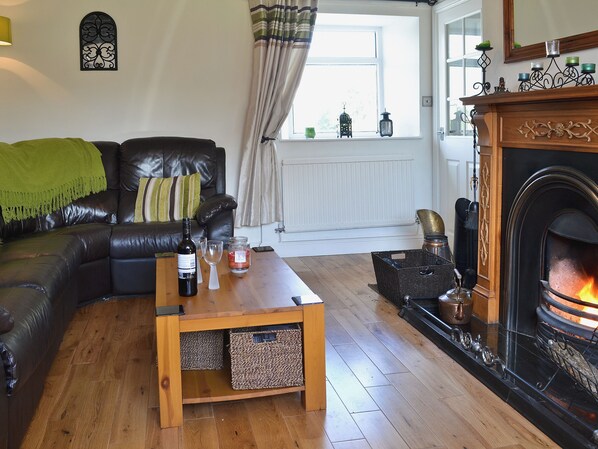 A cozy living room with a leather sofa, a wooden coffee table with a bottle and glasses, a fireplace, and a window with curtains. - 2 bedroom accommodation in Pen-y-sarn near Amlwch (Pen-y-sarn near Amlwch)