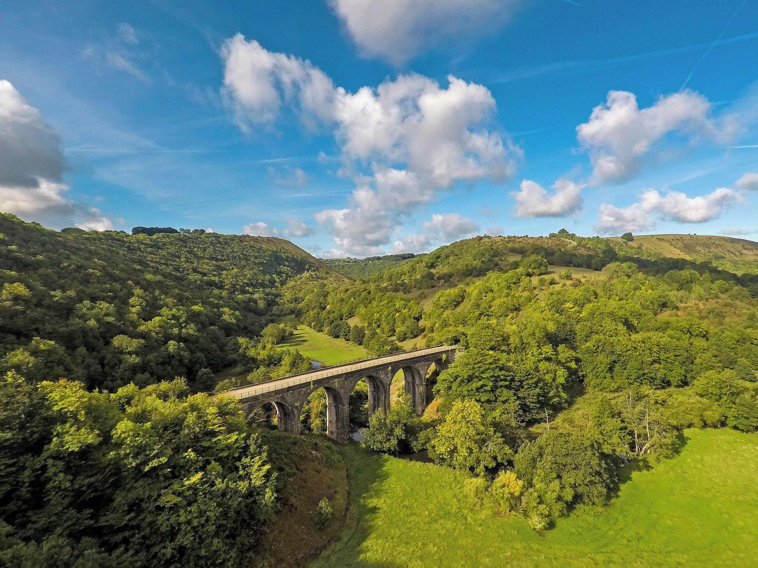 A stone bridge over a valley with lush greenery and hills in the background.