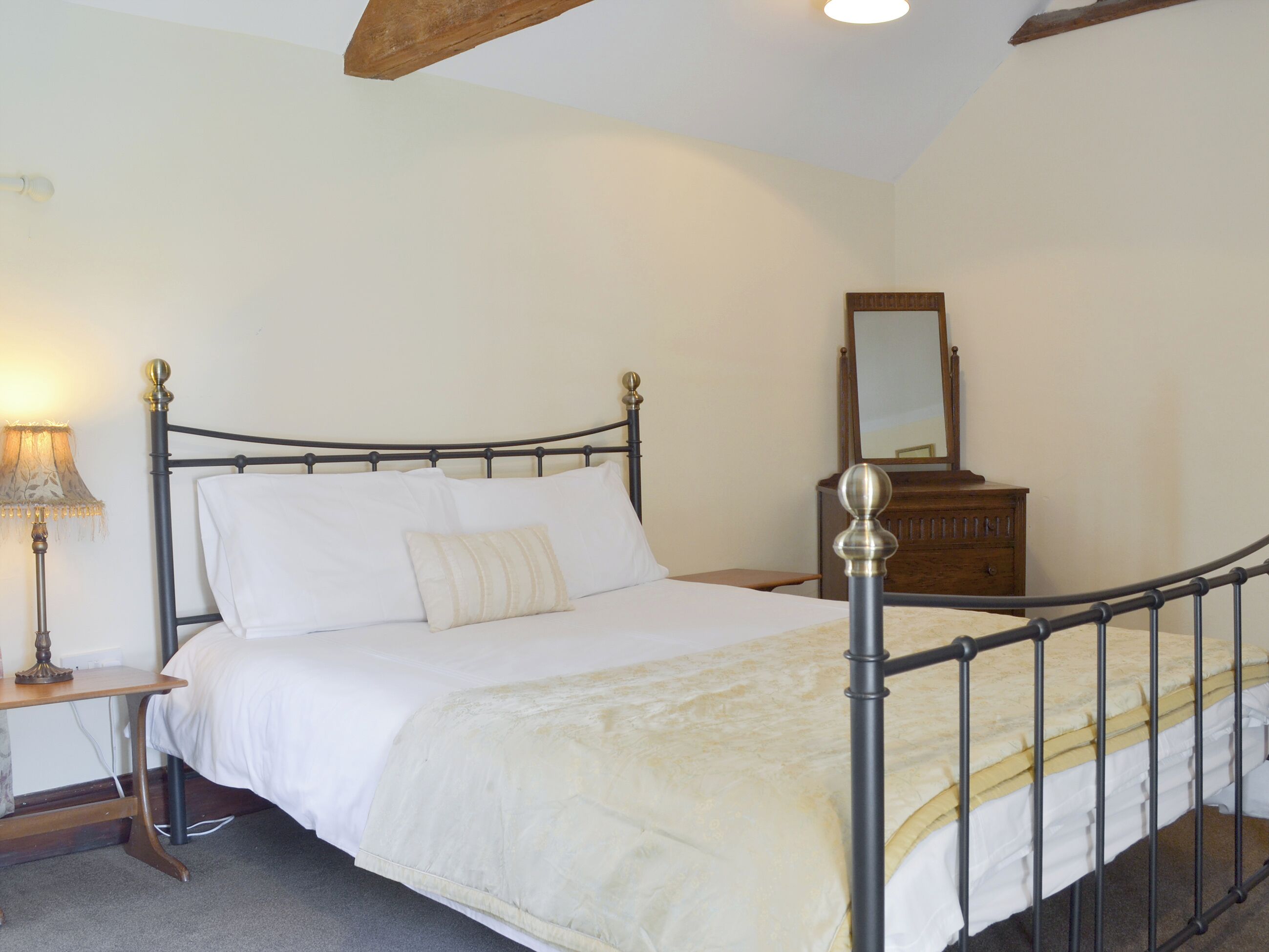 A neatly made bed with white linens and a single decorative pillow, a wooden nightstand with a lamp, and a wooden dresser with a mirror.