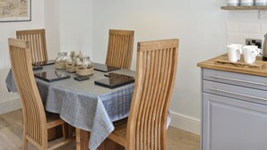 Zona pranzo con tavolo apparecchiato per quattro, circondato da sedie in legno, una lampada a sospensione bianca e angolo cottura con piano di lavoro e pensili in legno.