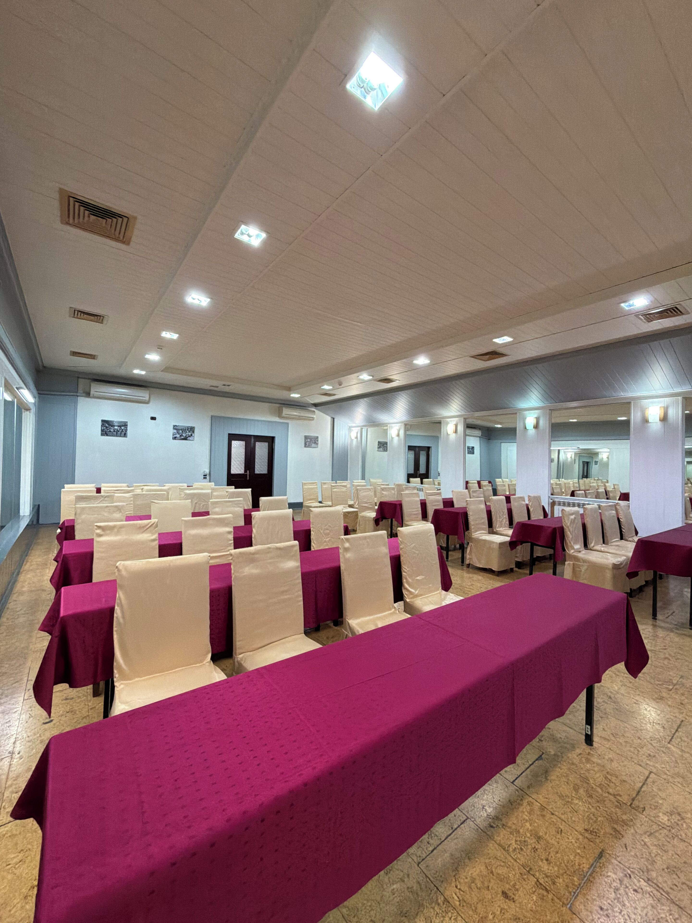 A conference room with rows of tables and chairs, air conditioning units, and a tiled floor.