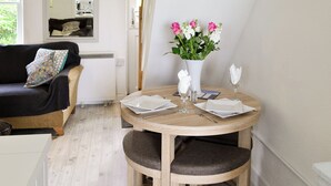 Een gezellige kamer met een ronde houten tafel gedekt voor twee personen, een witte vaas met bloemen, een bank met een decoratief kussen en een spiegel aan de muur.